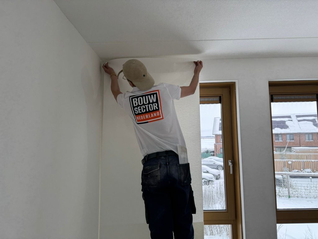 Behanger van Bouwsector Nederland drukt een baan renovlies bovenaan tegen de wand, vlak onder het plafond