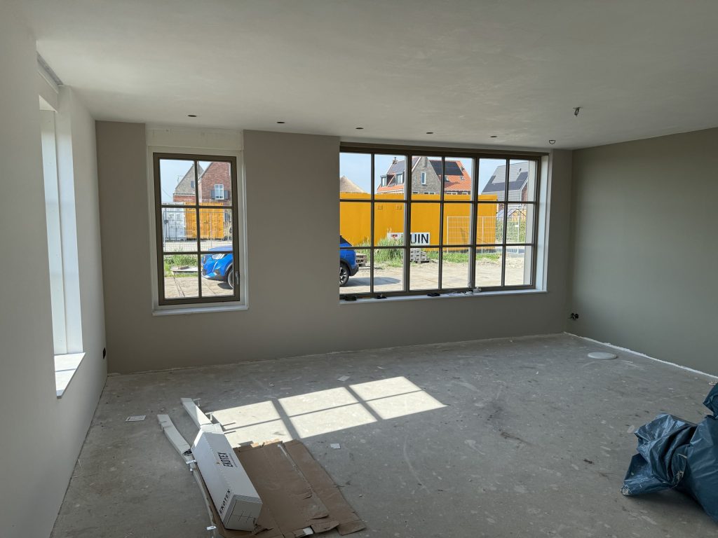 Lege woonkamer van een nieuwbouwwoning met grijsgroen gesausde wanden en ramen met zicht op huizen in aanbouw