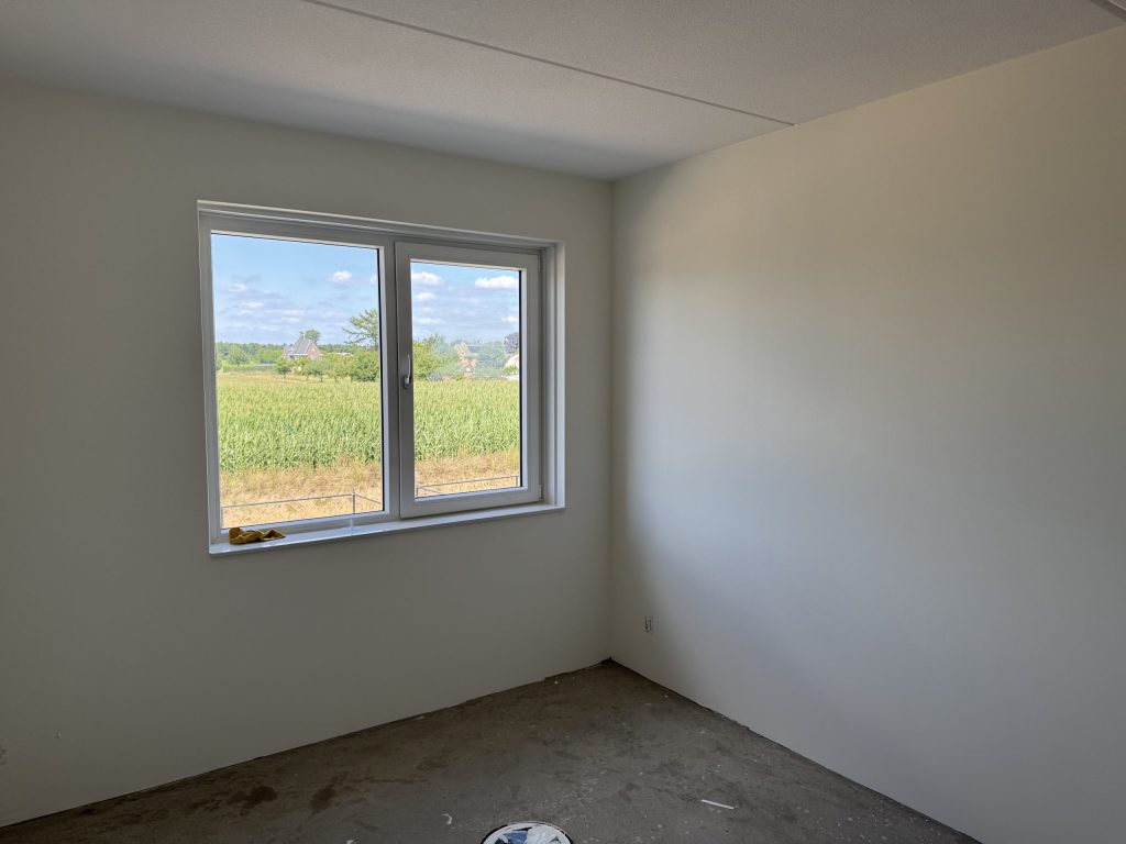 Lege slaapkamer in een nieuwbouwhuis met lichte wanden, een betonvloer en een raam dat uitkijkt op een akker