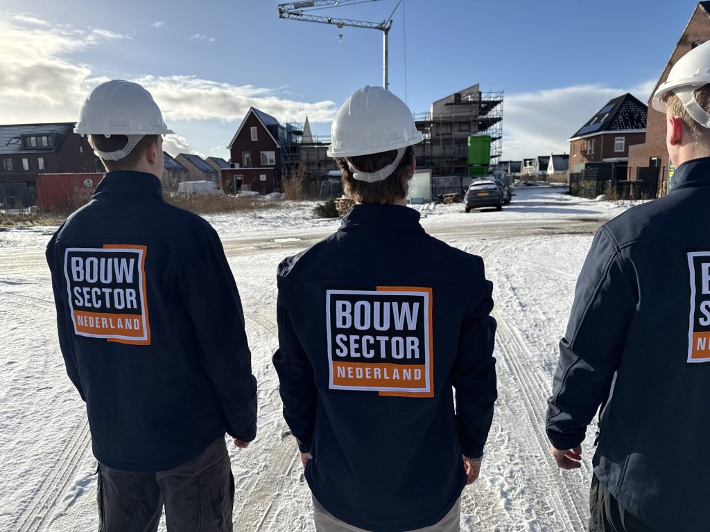 Drie vakmensen van Bouwsector Nederland met witte helm in een besneeuwde straat vol nieuwbouwhuizen en een bouwkraan